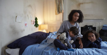 A mother and her young son and daughter sit on a bed in temporary accommodation that is in a bad state of disrepair