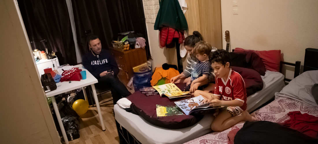 Father, mother and two young sons sit in a crowded bedroom in temporary accommodation