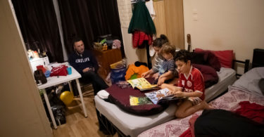 Father, mother and two young sons sit in a crowded bedroom in temporary accommodation