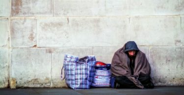 A homeless man sits on the ground with two bags filled with his belongings
