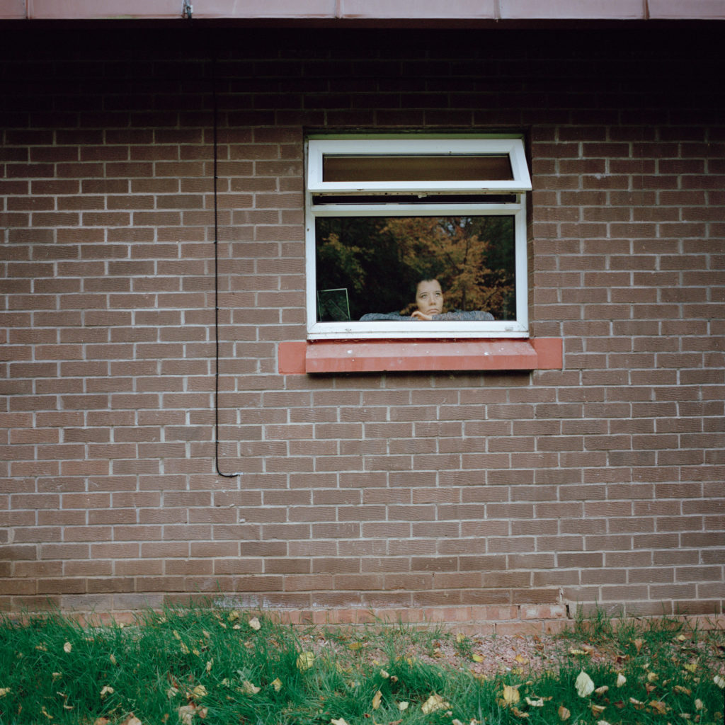A brown wall with a window in the middle. Lindsay is looking out with her chin reting on her arms. A tree is reflected in the window.