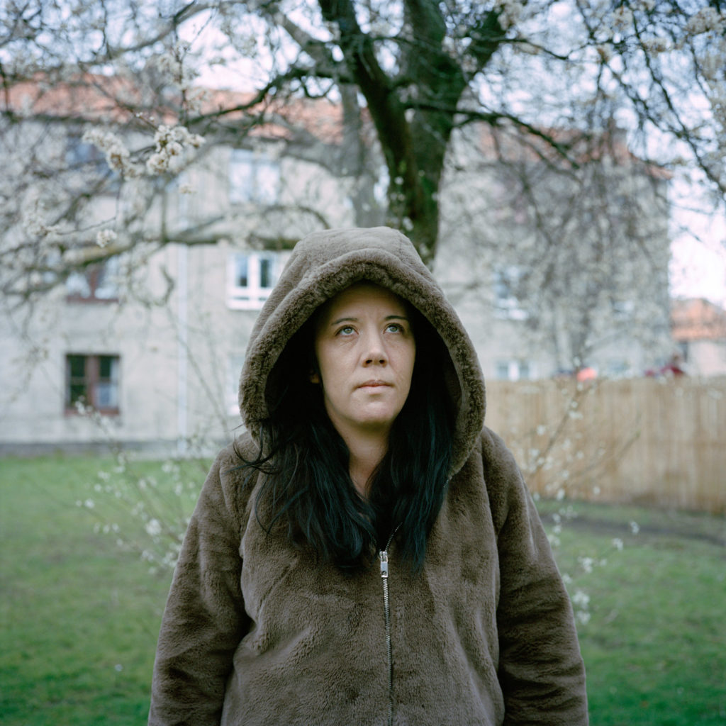 A portrait of Lindsay in a brown hoodie with the good up, standing in front of a tree and an apartment building.