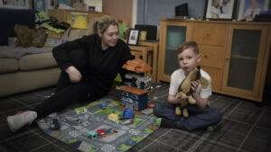 Stacey, wearing all black with blonde tied back hair, sitting on the floor smiling at her son Archie who his holding a toy dog.