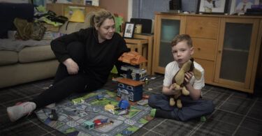 Stacey, wearing all black with blonde tied back hair, sitting on the floor smiling at her son Archie who his holding a toy dog.